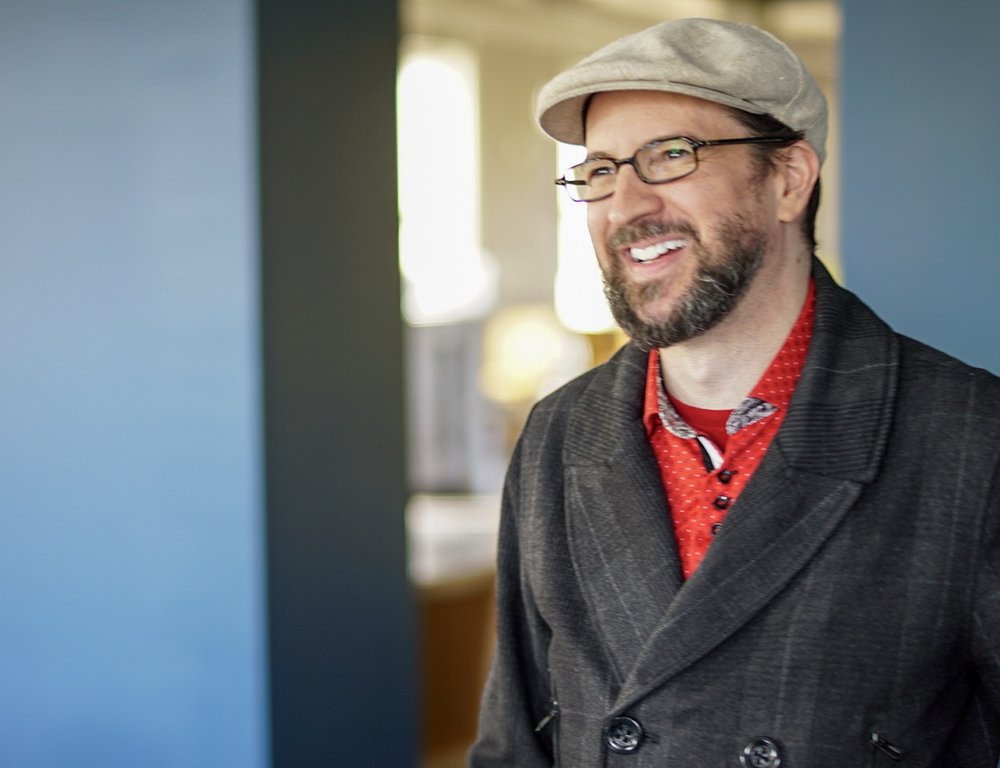 Photo of Kevin Brockmeier in red shirt, grey coat, glasses and tan newsboy cap